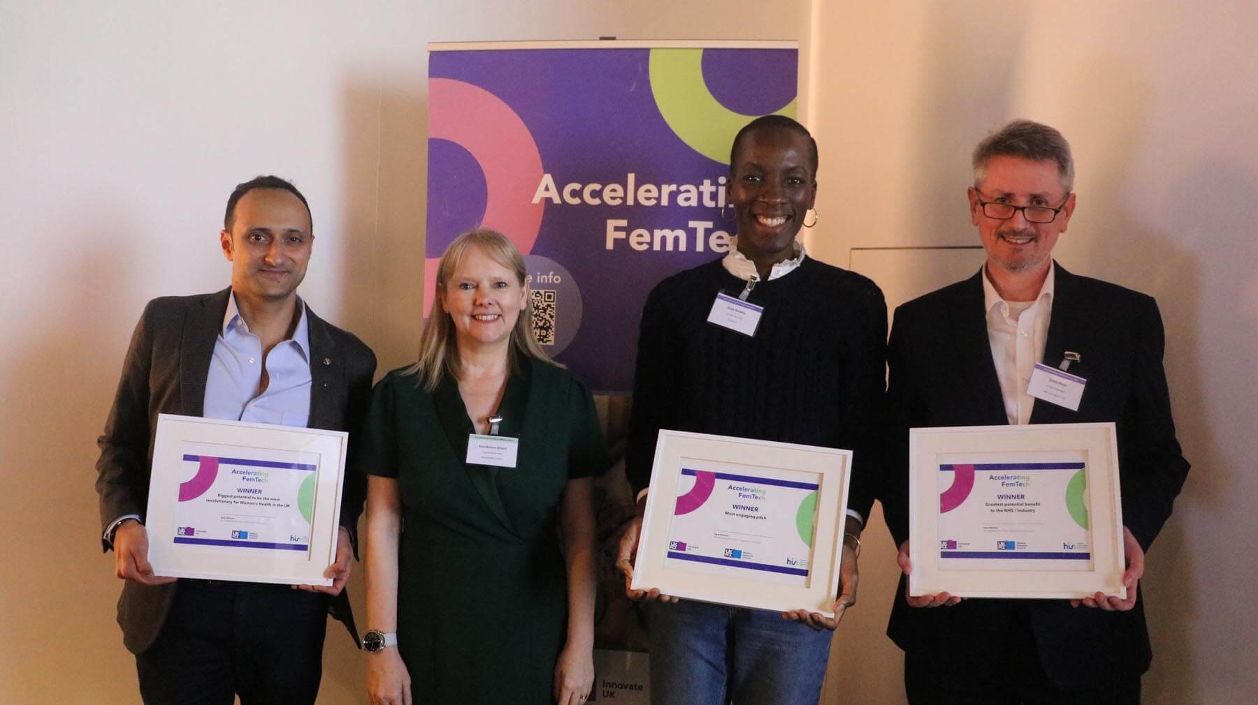 Four people stand in a row, smiling. Three people hold white frames with certificates. In the background, there is a banner with the text 'Accelerating FemTech'