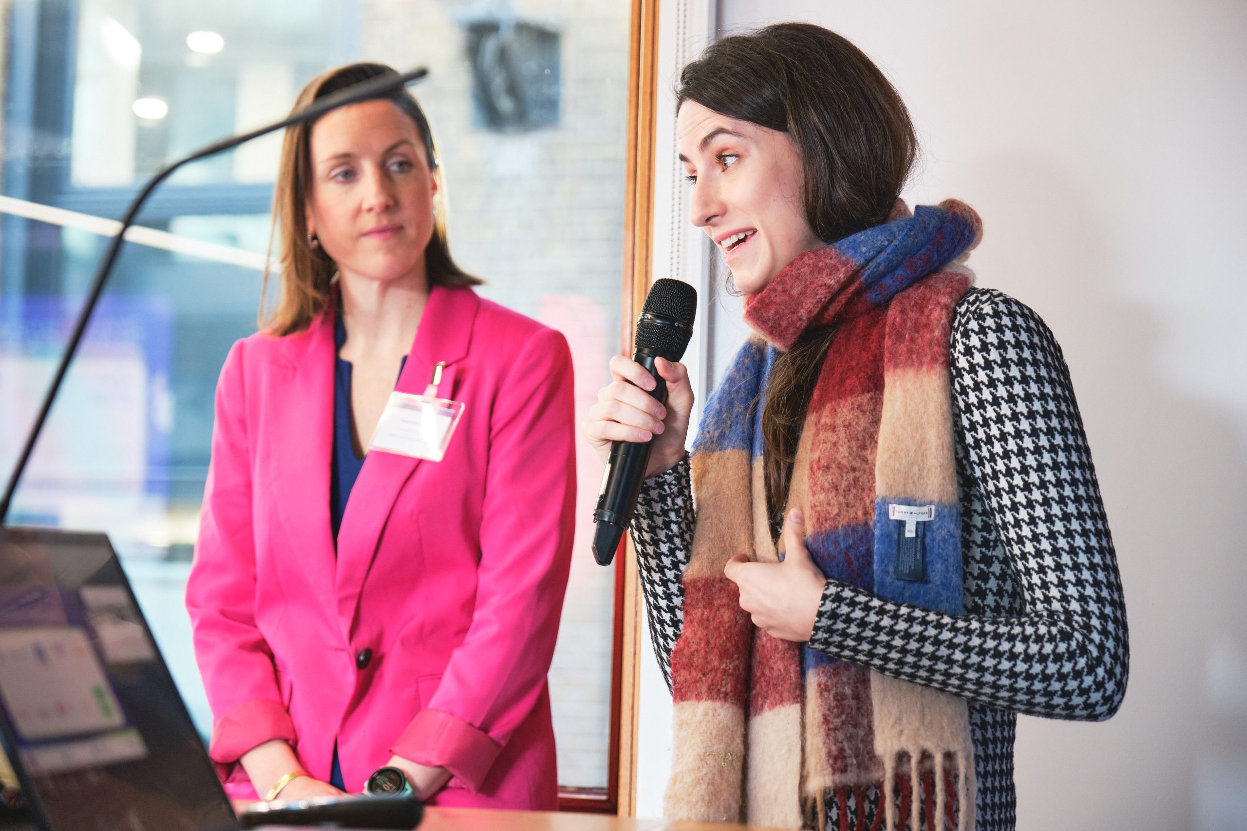 A person wearing a blue, red and cream scarf holds a microphone and speaks. In the background, a person wearing a pink blazer looks at the person speaking.